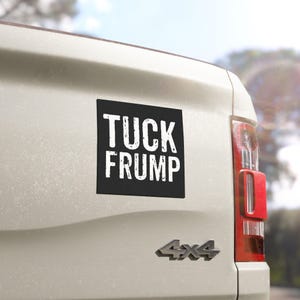 May include: A black square sticker with white text that reads "TUCK FRUMP" is attached to the back of a white pickup truck.