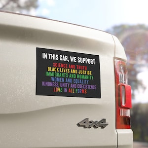May include: A rectangular black bumper sticker on a white vehicle. The sticker reads "IN THIS CAR, WE SUPPORT" followed by a list of values in rainbow colors: Science and Truth, Black Lives and Justice, Immigrants and Humanity, Women and Equality, Kindness, Unity and Coexistence, and Love in All Forms.