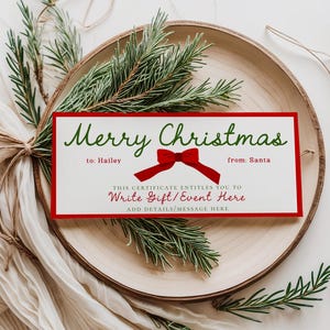 以下が含まれることがあります： 赤い縁と赤いリボンが付いたクリスマスギフト券。「Merry Christmas」の文字は緑色で書かれています。ギフト券は、ローズマリーの小枝が添えられた木製のプレートの上にあります。