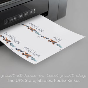 May include: A black printer outputting a white sheet of paper featuring printed labels with airplane illustrations and text including POPCORN, BEVERAGES, FUEL, and ROLL-UPS. The labels are arranged in rows with small blue and orange airplane graphics.