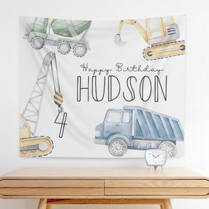 May include: A white fabric wall hanging featuring watercolor illustrations of construction vehicles: a cement mixer, excavator, crane, and dump truck. The text "Happy Birthday HUDSON" and the number "4" are also displayed.