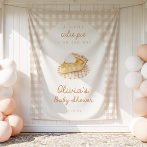 Puede incluir: Una pancarta de baby shower con un patrón de cuadros beige y blanco. La pancarta presenta el texto "A little cutie pie is on the way" y "Olivia's Baby Shower 11.16.20" con una ilustración de una porción de pastel. Globos blancos y melocotón decoran la escena.