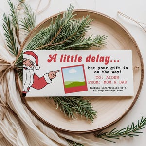 Puede incluir: Una tarjeta navideña rectangular con una ilustración de Papá Noel y el texto "A little delay... but your gift is on the way!". La tarjeta está sobre un plato de madera con vegetación y un paño blanco.