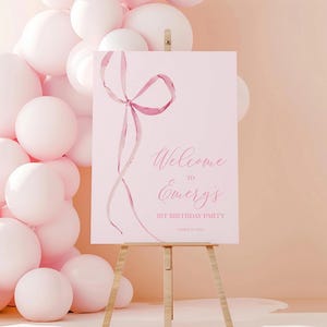 May include: Pink welcome sign with a pink ribbon bow and the text "Welcome to Emery's 1st Birthday Party".