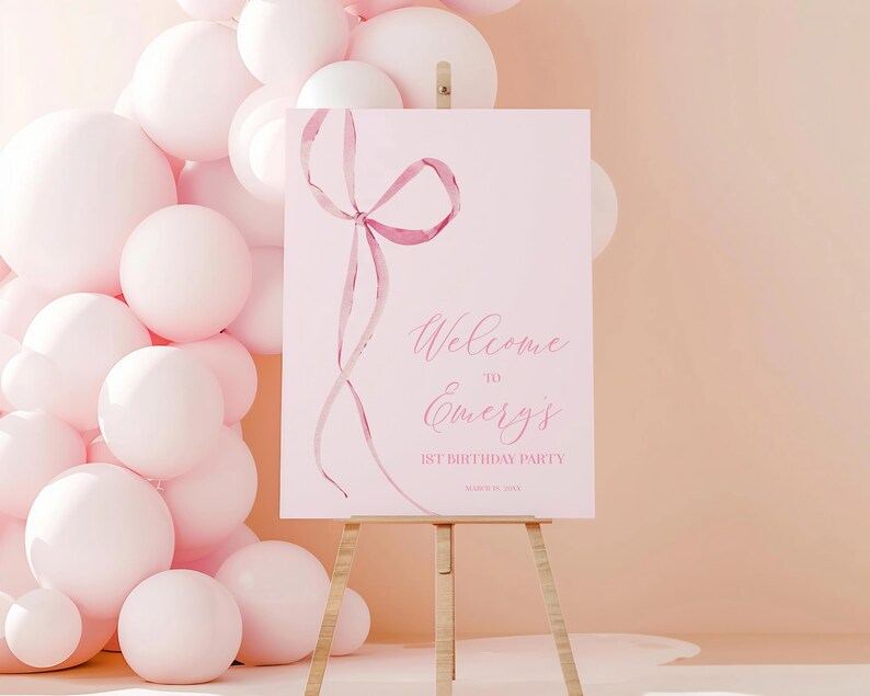 May include: Pink welcome sign with a pink ribbon bow and the text "Welcome to Emery's 1st Birthday Party".
