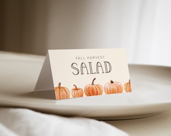 Op de afbeelding: Een witte kaart met de tekst "Fall Harvest Salad" in zwarte letters. De kaart is versierd met een rij van vijf kleine oranje pompoenen.