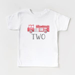 May include: A white short-sleeve t-shirt featuring a watercolor-style red fire truck graphic centered on the chest. Below the fire truck, the word TWO is printed in a bold, black, sans-serif font. The design is simple and centered on the shirt.