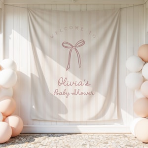 May include: A baby shower banner with the text "Welcome to Olivia's Baby Shower" in a soft pink colour, featuring a bow illustration. The banner is set against a striped background, with clusters of white and peach balloons.