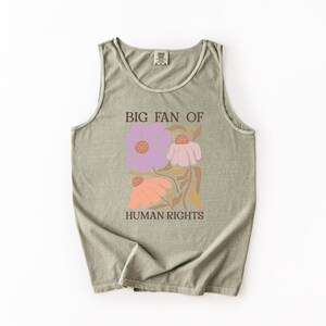 May include: A sage green sleeveless tank top featuring a graphic print of three stylized flowers in shades of purple, pink, and orange. The text "BIG FAN OF HUMAN RIGHTS" is printed in dark brown capital letters above and below the floral design.