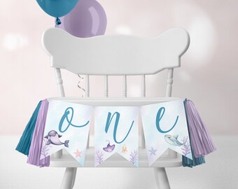 May include: A blue and purple high chair banner with the word "one" in blue cursive lettering. The banner features a variety of sea creatures, including a dolphin, a stingray, and an octopus.
