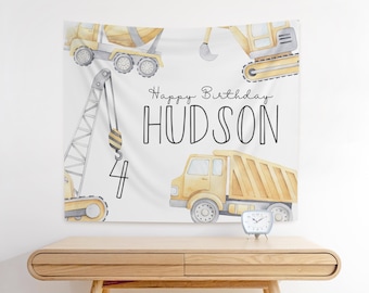 May include: A white banner with a construction theme for a birthday party. The banner features a yellow dump truck, a yellow crane, and a yellow cement mixer. The text on the banner reads "Happy Birthday Hudson".