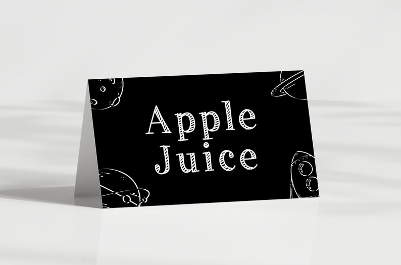 May include: Black and white sign with the text "Apple Juice" in a decorative font. The sign is decorated with white drawings of planets and spaceships.