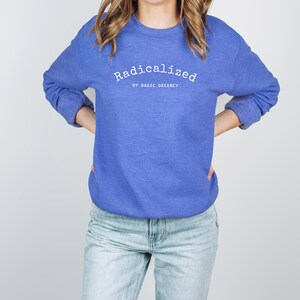 以下が含まれることがあります： 白字で「Radicalized」とプリントされた青いクルーネックのスウェットシャツ。下に「by Basic Decency」の文字。明るい青色のジーンズと合わせています。