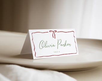 May include: A white place card with a red border and bow design. The name "Olivia Parker" is written in green script, with "TABLE 6" below. The card is on a white plate, with a white cloth in the foreground.