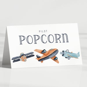May include: A white folded card featuring the text PILOT POPCORN in dark blue lettering. Below the text are three watercolor-style illustrations of airplanes: a dark blue biplane, an orange monoplane, and a light blue propeller airplane.