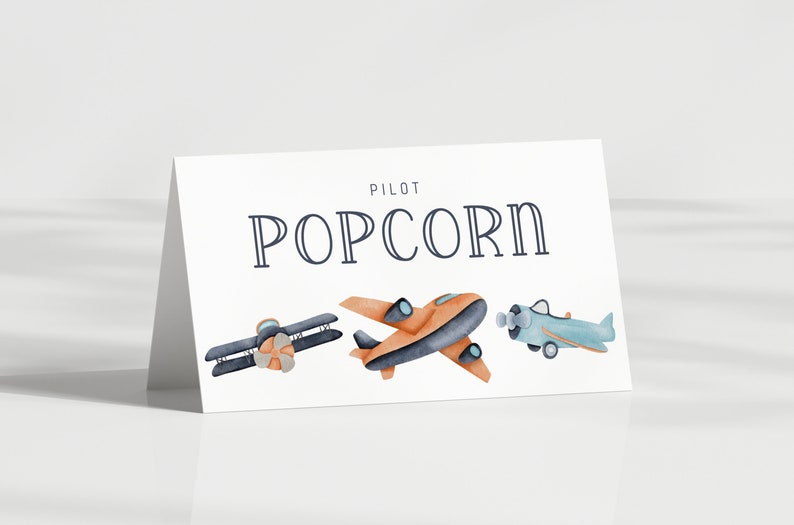 May include: A white folded card featuring the text PILOT POPCORN in dark blue lettering. Below the text are three watercolor-style illustrations of airplanes: a dark blue biplane, an orange monoplane, and a light blue propeller airplane.