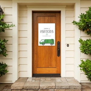 May include: Welcome to Ashton's 1st Birthday Party sign with a green garbage truck illustration. The sign is printed on white paper and is hanging on a wooden door.