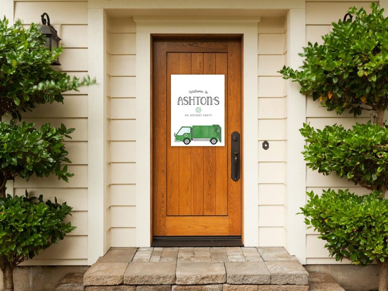 May include: Welcome to Ashton's 1st Birthday Party sign with a green garbage truck illustration. The sign is printed on white paper and is hanging on a wooden door.