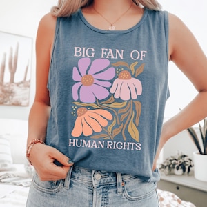 May include: A blue sleeveless tank top featuring a graphic print with the text "BIG FAN OF HUMAN RIGHTS" in light pink. The design includes three stylized flowers in shades of purple, light pink, and orange, accompanied by green foliage.