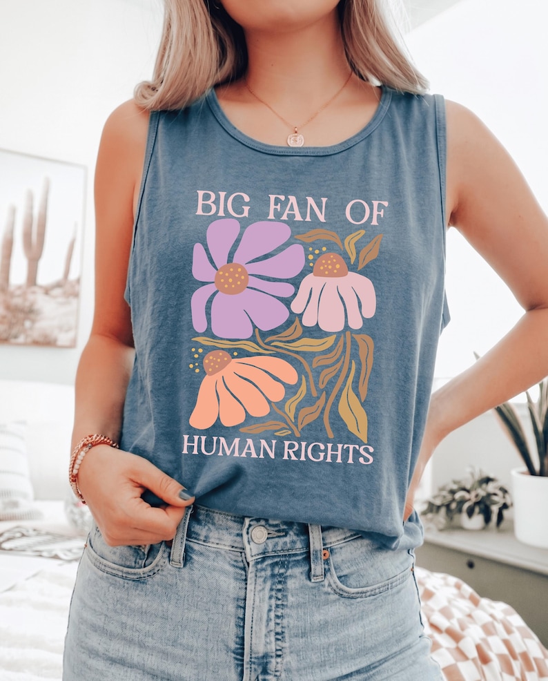 May include: A blue sleeveless tank top featuring a graphic print with the text "BIG FAN OF HUMAN RIGHTS" in light pink. The design includes three stylized flowers in shades of purple, light pink, and orange, accompanied by green foliage.
