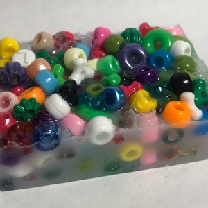 May include: A clear block of resin filled with colorful plastic beads. The beads are in various shapes, including circles, squares, and stars. The resin is clear and allows the beads to be seen clearly.