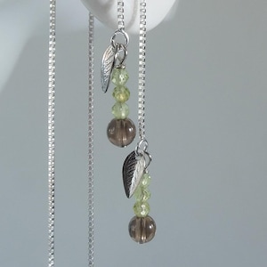 以下が含まれることがあります： 葉のチャーム、緑色のビーズ、茶色のビーズが付いたシルバーチェーンのイヤリング。白い耳からぶら下がっているイヤリングが写っています。繊細でエレガントなデザインのイヤリングです。