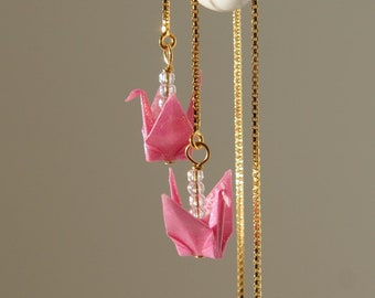 May include: Pink origami crane earrings with gold-colored chains and small clear beads. The earrings are hanging from a white surface. The earrings are a unique and delicate accessory.