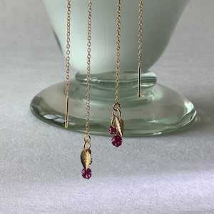 以下が含まれることがあります： 葉の形をしたチャームと小さな丸い赤い宝石が付いた、金色のスレッドピアス。白いガラス製の装飾花瓶の前にイヤリングがぶら下がっています。