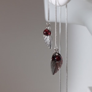 以下が含まれることがあります： 小さな赤い宝石がベースについた、銀色の葉のイヤリング。イヤリングは繊細な銀色のチェーンから吊り下げられています。イヤリングは白いマネキンの頭に展示されています。