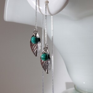 以下が含まれることがあります： 葉のデザインのシルバーカラーのぶら下がりイヤリング。それぞれに小さな丸いターコイズ色の石が付いています。イヤリングは繊細なチェーンから吊り下げられ、白いマネキンの頭に展示されています。ジュエリーアイテムです。