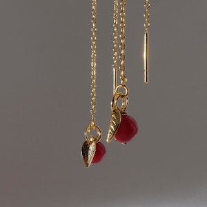 以下が含まれることがあります： 赤いビーズと葉のチャームが付いたゴールドカラーのチェーンピアス。ピアスは繊細なデザインで、チェーンが垂れ下がっています。赤いビーズは丸く、葉のチャームは金色です。ピアスはニュートラルな背景に表示されています。