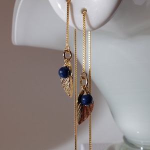 以下が含まれることがあります： 金色のチェーンピアス。葉のチャームと濃い青色のビーズが付いています。ピアスは白い表面から吊り下げられています。葉のチャームには複雑なディテールがあります。ピアスは繊細でエレガントです。