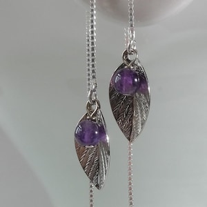以下が含まれることがあります： 小さな紫色の宝石が付いた、銀色の葉のイヤリング。イヤリングは繊細なチェーンから吊り下げられています。白い装飾品に展示されています。「PrincessTreeCraft」のロゴが左下に表示されています。
