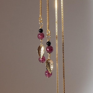 以下が含まれることがあります： ゴールドトーンのチェーンピアス。葉のチャーム、ピンクと黒のビーズが揺れるデザイン。白いマネキンの頭に展示されています。繊細でエレガントなデザインのピアスです。