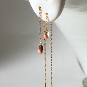 以下が含まれることがあります： 小さな桃色の涙型のビーズが付いた、ゴールドカラーのチェーンピアス。 ピアスは、各チェーンの端に垂直なバーが付いた、長く垂れ下がるデザインです。 白い背景に表示されています。