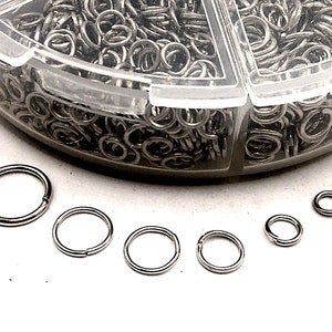 May include: A clear plastic container filled with silver jump rings of various sizes. The container has six compartments. There are five silver jump rings of different sizes on a white surface.