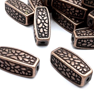May include: A pile of copper-colored metal beads with a textured, patterned surface. The beads are oval-shaped and have a hole through the center.