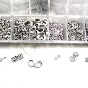 May include: A clear plastic storage box with multiple compartments filled with silver metal beads, charms, and spacers. The box is open and some of the beads are scattered on the white surface below.