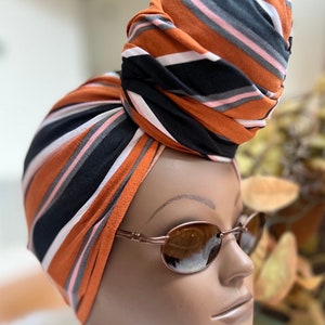 May include: A brown, black, and pink striped turban with a knot on top. The turban is worn on a mannequin head with brown oval sunglasses.
