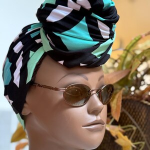 May include: A mannequin head wearing a black, teal, and white patterned turban and brown oval sunglasses. The turban is wrapped in a stylish design, and the sunglasses have a thin, gold-colored frame.