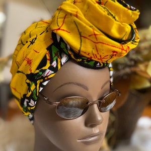 May include: A yellow and green patterned head wrap with a black trim. The wrap is tied in a turban style. A person is wearing the wrap and brown oval sunglasses.