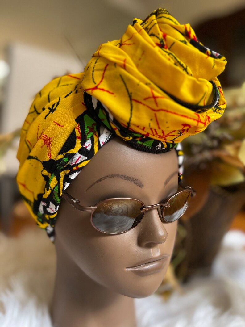 May include: A yellow and green patterned head wrap with a black trim. The wrap is tied in a turban style. A person is wearing the wrap and brown oval sunglasses.