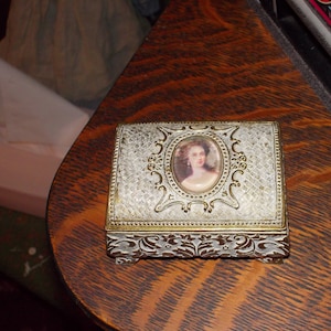 May include: A small, ornate, silver-colored trinket box with a white and gold design. The box has a raised, oval-shaped image of a woman with dark hair and a white dress. The box is sitting on a wooden surface.