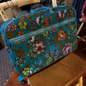 May include: A rectangular suitcase featuring a vibrant teal background with a repeating floral pattern in shades of orange, purple, yellow, and green. The luggage includes a top handle, a zippered front pocket, and a main zipper closure along the top.