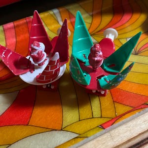 May include: Two Christmas-themed ornaments. One is red and white, featuring a Santa Claus figure and a chimney. The other is green and red, also with a Santa Claus figure. Both ornaments have a unique, stylized design, perfect for holiday decor.