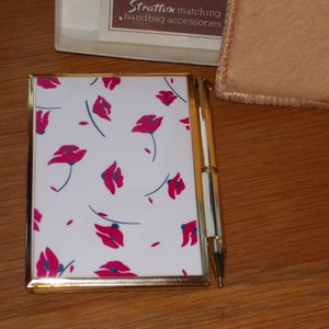 May include: A white notepad with a gold frame and a red poppy floral pattern. The notepad is closed and has a gold pen next to it.