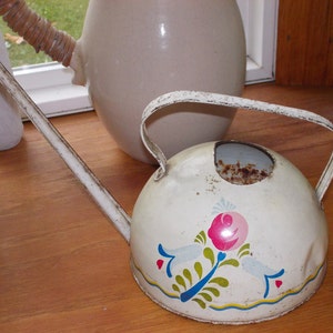 May include: A vintage, white metal watering can with a long spout and a curved handle. The can features a hand-painted floral design in pink, blue, and yellow. The watering can is sitting on a wooden surface.