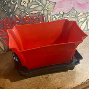 May include: A red plastic planter with a decorative, scalloped edge sits on top of a black plastic tray. The planter and tray are both empty.