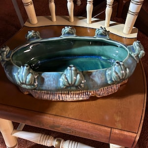 May include: An oval, ceramic centerpiece bowl in shades of teal and blue, adorned with frog figurines around the rim. The bowl sits on a wooden chair with a brown seat and white legs.
