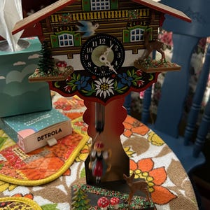 May include: A wooden cuckoo clock with a red roof and a white face. The clock has a blue bird, a deer, and a girl on a swing. The clock is decorated with flowers and mushrooms.
